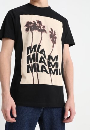 Camiseta de algodón negra con un gran gráfico de palmeras y la palabra "MIAMI" en texto negro en negrita sobre un fondo beige.
