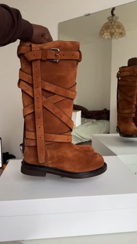 Barna velúr térd fölé érő csizma keresztben átkötött pántokkal és csattal díszítve. Vastag talp varrott mintával, sima textúrával és kerek orral.