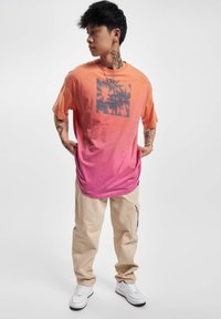 Kortermet gradient t-skjorte, fra oransje til rosa, med et mørkt palmetre-grafikk. Kombinert med beige cargo-bukser og hvite sneakers.