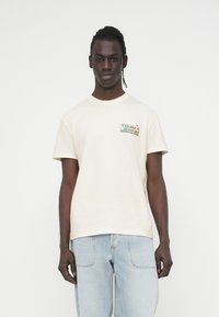 Μπεζ β αμβακερό t-shirt με μικρό, έγχρωμο λογότυπο "TOMMY JEANS" στο στήθος. Χαλαρή εφαρμογή με κοντά μανίκια και κλασικό στρογγυλό λαιμό.