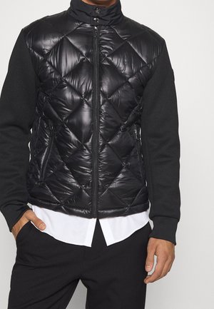 Homme portant une veste matelassée noire zippée avec des manches en tricot par-dessus une chemise blanche, debout avec une main dans la poche.
