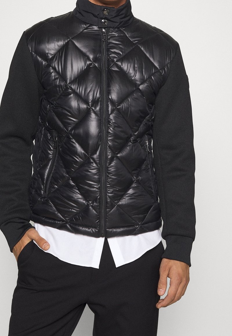 Homme portant une veste matelassée noire zippée avec des manches en tricot par-dessus une chemise blanche, debout avec une main dans la poche.