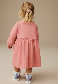 Rosa, transparente Kleid mit geraffter Taille und langen Ärmeln. Akzentuiert durch weiße Spitze an den Bündchen und bunten, gemusterten Turnschuhen.