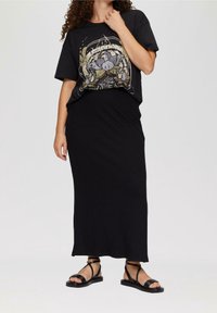 T-shirt noir en coton avec un design graphique, associé à une jupe longue à côtes noires et des sandales à lanières noires, présentant un look monochrome.