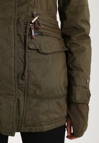 Detailní záběr na kapsu zelené prošívané bundy se zipem a šňůrkou, která je nošena s odpovídajícími pletenými rukavicemi bez prstů a černými kalhotami.