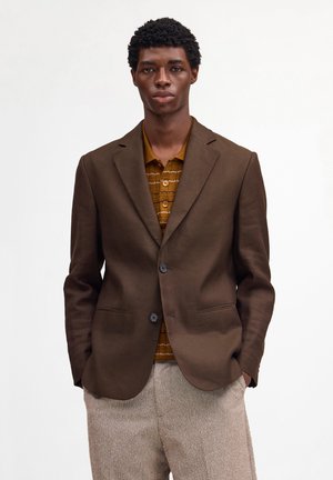 Mand iført brun blazer, sennepsfarvet strikket poloshirt og beige teksturerede bukser, stående med hænderne i lommerne mod en hvid baggrund.