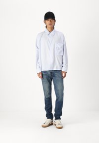Light blue striped button-up shirt with a chest pocket, paired with faded blue jeans and white shoes with beige soles. Black cap worn.