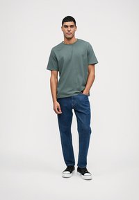 Σκούρο πράσινο βαμβακερό T-shirt με στρογγυλή λαιμόκοψη, συνδυασμένο με μπλε τζιν παντελόνι και μαύρα αθλητικά παπούτσια με λευκές σόλες. Στάση όρθια.