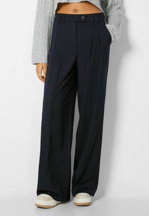 Woman wearing black pinstripe wide-leg trousers, gray cropped sweater, and white platform shoes, standing with one hand in pocket.