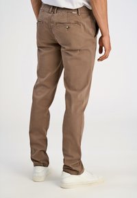 Uomo che indossa pantaloni casual marroni e sneakers bianche, in piedi con le mani rilassate ai lati su uno sfondo chiaro uniforme.