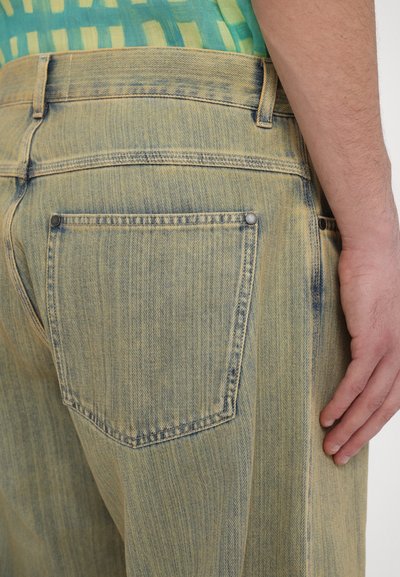 Faded yellow denim jeans shown from the back, featuring a back pocket with a rivet and visible stitching along the waistband and seams.
