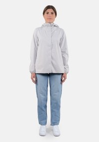 Giacca impermeabile grigia chiara con cappuccio, polsini elasticizzati e due tasche laterali, abbinata a jeans in denim azzurro chiaro e sneakers bianche.