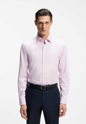 Uomo con capelli tirati indietro che indossa una camicia elegante rosa chiaro infilata in pantaloni scuri con cintura nera.