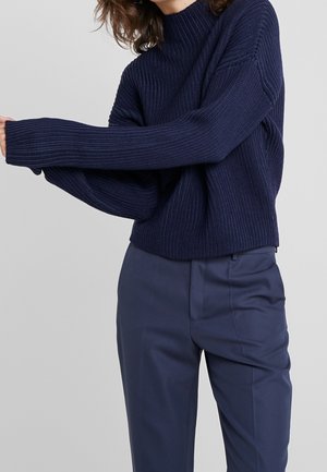 Personne portant un pull à col roulé côtelé bleu marine à manches longues et un pantalon bleu marine tailleur assorti.