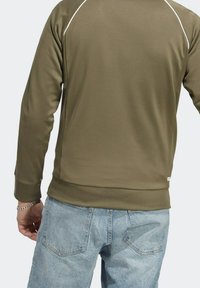 Oliwkowo zielony pulower o gładkiej fakturze, z białym ozdobnym przeszyciem i ściągaczami na mankietach, zestawiony z jasnoniebieskimi dżinsami.