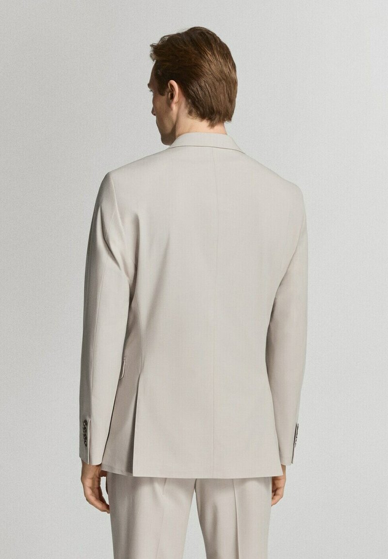 Hombre con cabello castaño vistiendo una chaqueta de traje beige claro y pantalones a juego, visto desde atrás sobre un fondo liso.