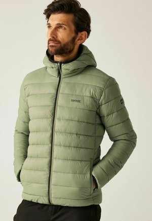 Hombre con barba que lleva una chaqueta acolchada con capucha de color verde claro, con cremallera y manos en los bolsillos, mirando hacia la izquierda sobre un fondo liso.