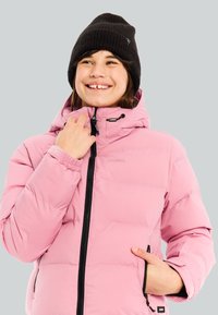 Persona sonriente que lleva un gorro de lana negro y una chaqueta de plumas rosa, sosteniendo la cremallera con una mano y un bolsillo con la otra.