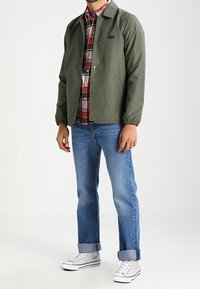 Giacca leggera verde, camicia a quadri, jeans blu e sneakers bianche. La giacca presenta polsini elastici e un piccolo logo.