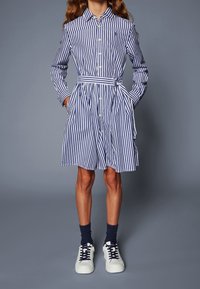 Dívka v modro-bíle pruhované šatech s opaskem a kapsami, v námořnických ponožkách a bílých teniskách, stojí na šedém pozadí.