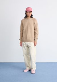 Beige gestrickter Pullover mit rundem Halsausschnitt, weißen weiten Hosen und einer rosa Baseballmütze. Hellrosa Sneakers runden das Outfit ab.