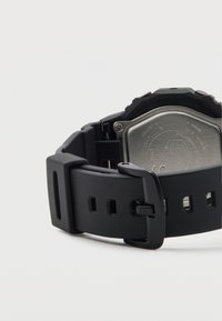 Czarny cyfrowy zegarek z gumowym paskiem, stalowym tyłem i regulowaną klamrą. Posiada kwadratową kopertę i widoczne otwory na pasku do regulacji rozmiaru.