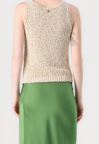 Top sin mangas de textura beige con escote en la espalda y un pequeño detalle de logo, combinado con una falda de satén verde ajustada.