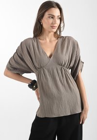 Blusa marrone a trama con scollo a V profondo, maniche corte a kimono e vita arricciata. Indossata con pantaloni neri e un bracciale spesso.