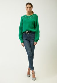 Grüner gestrickter Pullover mit dekorativen Perlenverzierungen am Ausschnitt, kombiniert mit dunklen Skinny-Jeans und silbernen High Heels.
