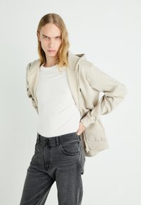 Sudadera con cremallera de color beige claro, con un bolsillo frontal, llevada sobre una camiseta blanca. Combinada con jeans gris oscuro de corte clásico y con bolsillos.