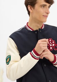Marineblaues und cremefarbenes Varsity-Jacket mit roten Akzenten, ausgestattet mit einem strukturierten Logo auf der Brust und einem gestickten Patch am Ärmel.