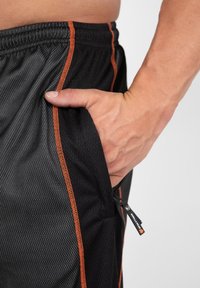 Zwarte sportshorts met een oranje zijstreep, gemaakt van een gestructureerd mesh-materiaal en voorzien van een zijzak. Een kleine ritssluiting is zichtbaar.