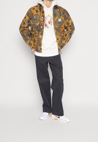 Ruskea kuviollinen takki valkoisen hupparin päällä, yhdistettynä tummaan housuun ja valkoisiin lenkkareihin. Takissa on paisley-kuvioita ja napit.