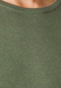 Olijfgroene gebreide trui met een geribde halslijn en een gladde textuur, met een close-up van het geweven patroon van de stof.