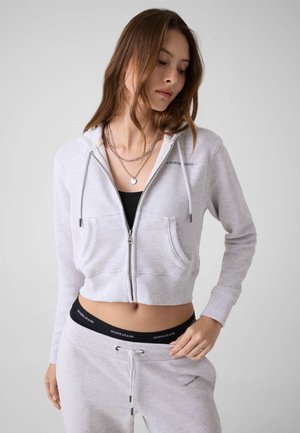 Femme portant un sweat à capuche zippé gris clair, un pantalon de jogging assorti et des colliers superposés, debout avec une main sur la hanche, regardant vers le bas.