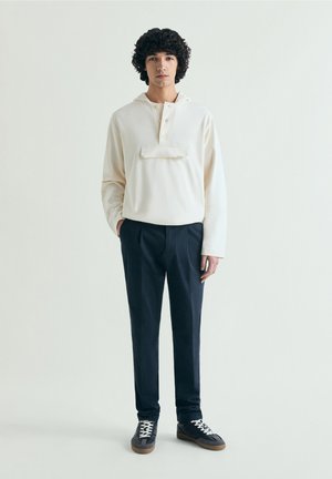 Joven con pelo rizado que lleva una sudadera con capucha cremosa, pantalones azul marino y zapatillas negras con cordones blancos, de pie frente a un fondo liso.