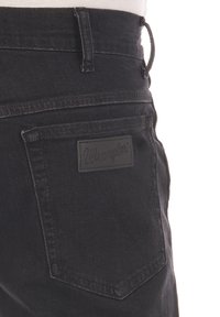 Jeans de mezclilla negros con diseño estructurado, bolsillo trasero, detalles con costuras y una parche rectangular de cuero negro con la inscripción "Wrangler" en relieve.