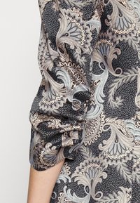 Blusa a maniche lunghe in tessuto nero con intricata stampa paisley blu e beige. Presenta polsini arricciati e una texture liscia.
