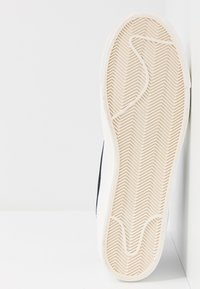 White sneaker sole with herringbone tread pattern, featuring a black accent on the side. Smooth rubber material and stitched edge detail.