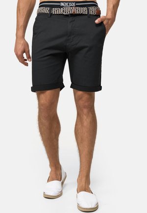 Homme portant un short noir retroussé avec ceinture tissée, des chaussures blanches sans lacets et la ceinture visible d'un sous-vêtement INDICODE.