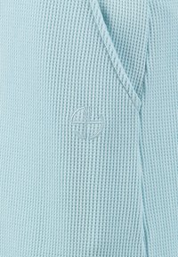 Tessuto a maglia a nido d'ape azzurro chiaro con un sottile logo circolare ricamato vicino a un bordo piegato.