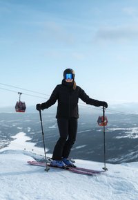 Personne en tenue de ski noire et casque, debout sur des skis sur une pente enneigée avec des bâtons de ski, montagne et télécabines en arrière-plan.