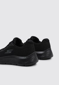 Czarne buty sportowe z siateczkową cholewką, wyściełanym kołnierzem oraz teksturowaną czarną podeszwą z oznakowaniem 'Go Walk' po boku.