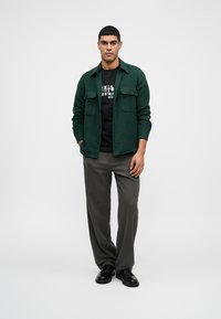 Muž oblečený v tmavozelenej zapínacej bunde, čiernom tričku s potlačou, šedých voľných nohaviciach a čiernych topánkach, stojí s rukami v vreckách.