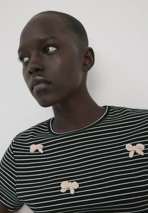 Mladá osoba s tmavou pokožkou a krátkymi vlasmi, ktorá má na sebe čierno-biele pruhované tričko s tromi malými béžovými pletenými mašličkami.