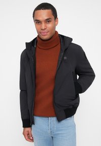 Veste à capuche noire avec fermeture éclair, présentant un extérieur lisse et des poignets côtelés. Associée à un pull col roulé côtelé orange foncé.