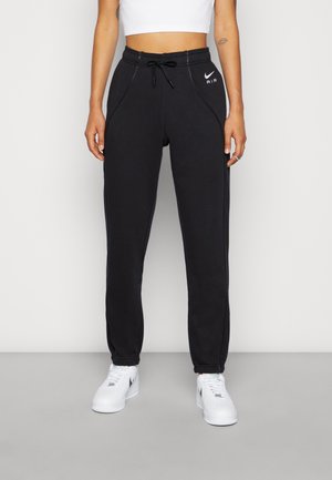 Kvinde iført sorte Nike Air joggers med snøre i taljen, kombineret med hvid crop top og hvide Nike sneakers, stående mod en ensfarvet lys baggrund.