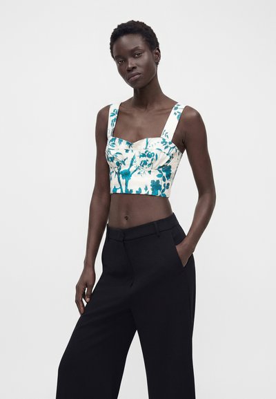 Femme portant un crop top blanc à imprimé floral bleu et un pantalon noir taille haute à jambes larges, debout avec une main dans la poche.