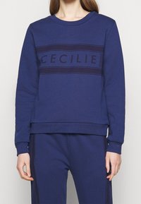 Námořnická modrá mikina s kulatým výstřihem, žebrovanými manžetami a lemem. Obsahuje horizontální pruh s textem "CECILIE" v tmavším odstínu.