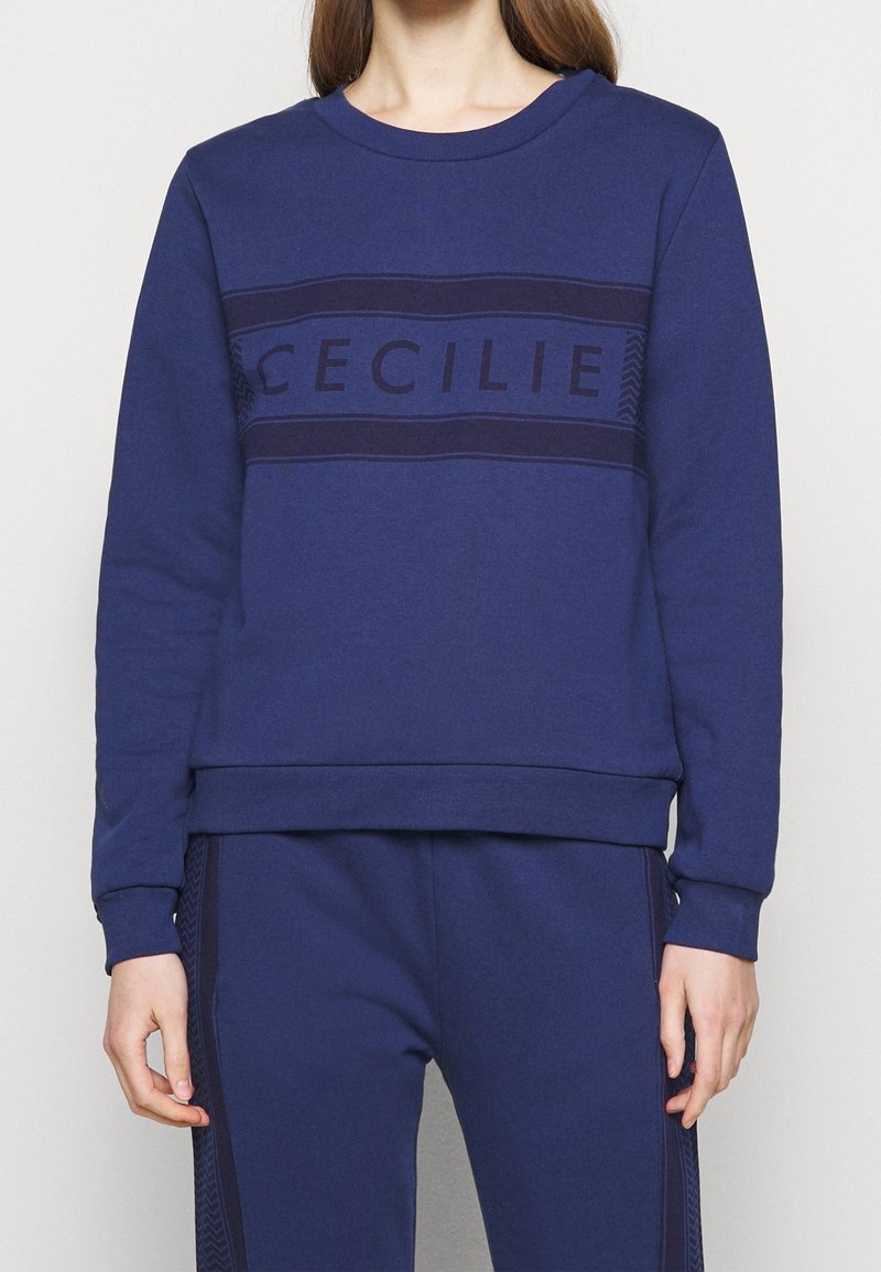 Námořnická modrá mikina s kulatým výstřihem, žebrovanými manžetami a lemem. Obsahuje horizontální pruh s textem "CECILIE" v tmavším odstínu.